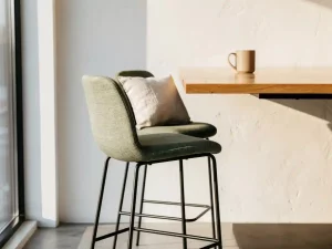 Twee gestoffeerde barkrukken onder een minimalistische eikenhouten hoge tafel in een lichte moderne kantoorhoek met linnen kussen en keramische mok.