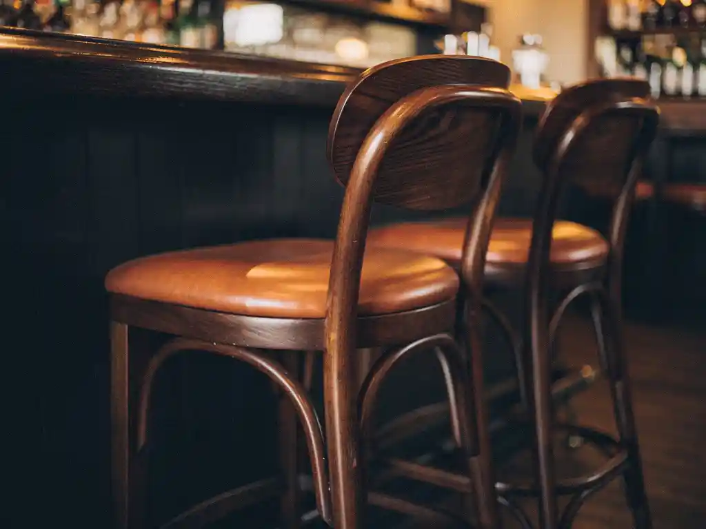 Drie houten barkrukken met lederen zitting langs een donkere eiken bartafel in een sfeervolle bar met warm amberkleurig licht.