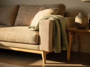 Linnen bank met diepe kussens en houten poten in zonnige woonkamerhoek, wollen plaid over armsteun, keramische vaas op bijzettafel.