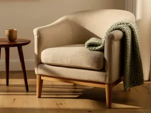 Linnen fauteuil met houten poten en wollen plaid in warme woonkamerhoek met keramieken kopje en zacht middaglicht.