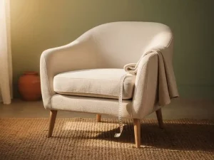 Linnen fauteuil met houten poten en meetlint op de armleuning, op een juten vloerkleed in een woonkamer met saliegroen muur.