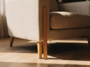 Linnen fauteuil in zandtint met houten meetlint langs de poot voor zithoogte, warm middaglicht op eiken vloer.