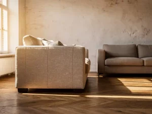 Op maat gestoffeerde linnen bank naast showroombank op visgraateikenhouten vloer in zonnig Nederlands interieur.